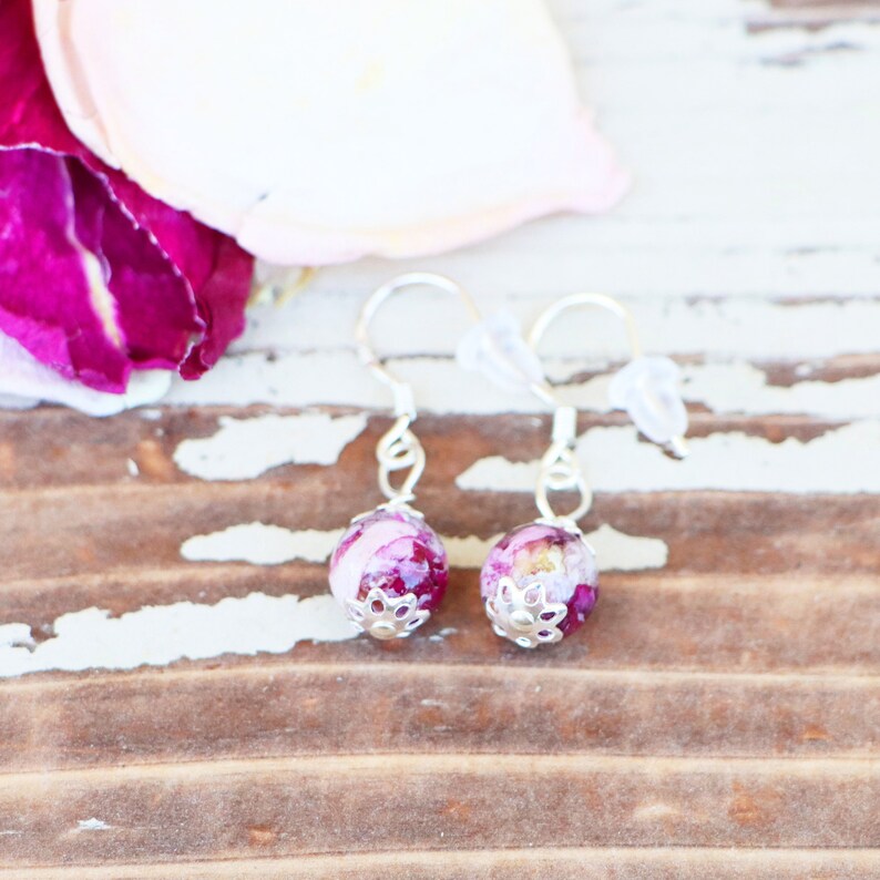May include: A pair of silver earrings with pink and white flower petals encased in clear resin. The earrings have a delicate, floral design.