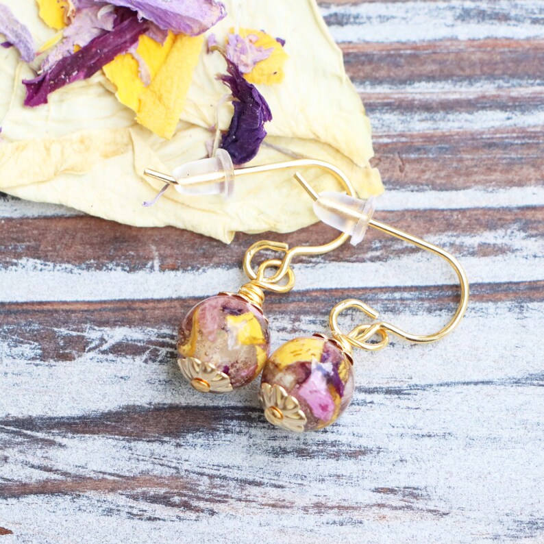May include: A pair of gold earrings with small round beads filled with pressed flowers in shades of pink, yellow, and purple.
