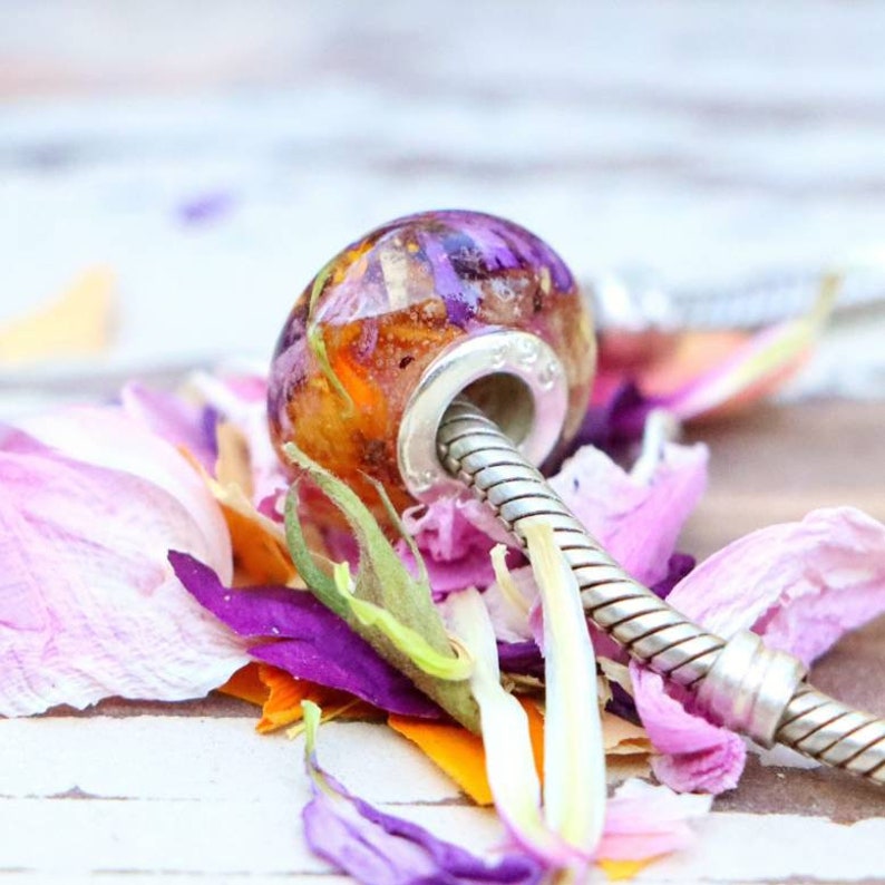 May include: A clear, round bead with a silver-colored core, filled with dried purple and orange flowers. The bead is resting on a bed of purple, pink, and orange flower petals, with a silver-colored bracelet chain.