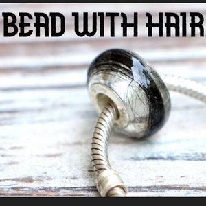 May include: A bead with the text "BEAD WITH HAIR" is shown. The bead is oval-shaped, with a silver-colored metal base and a clear, glossy top encasing black hair strands. It is attached to a silver-colored, flexible bracelet chain.