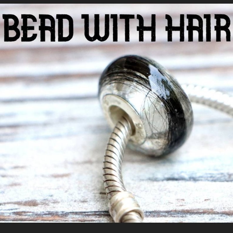 May include: A bead with the text "BEAD WITH HAIR" is shown. The bead is oval-shaped, with a silver-colored metal base and a clear, glossy top encasing black hair strands. It is attached to a silver-colored, flexible bracelet chain.
