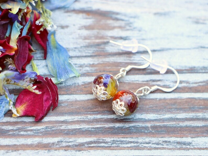 May include: A pair of silver earrings with round, colorful resin beads. The beads are filled with pressed flowers and have a silver filigree design around the edge.