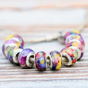 May include: A bracelet featuring colorful glass beads with floral designs. The beads are round with silver-colored metal accents. The flowers are in shades of purple, yellow, and red.
