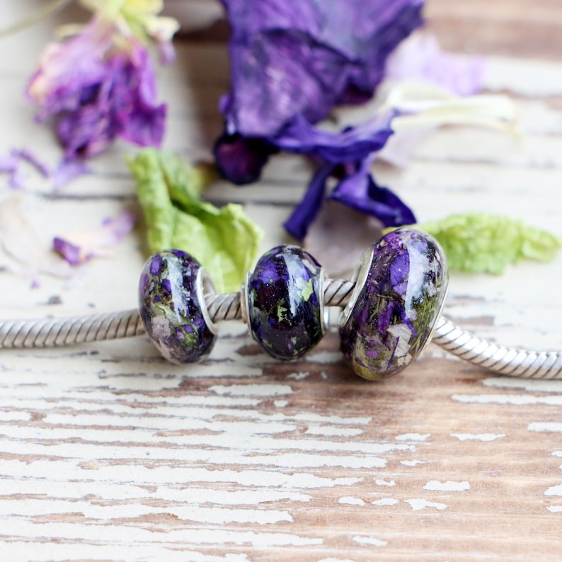 May include: Three glass beads with silver-colored metal cores strung on a silver chain. The beads are filled with purple and green dried flowers. The background includes purple flower petals and green leaves on a white, distressed wooden surface.