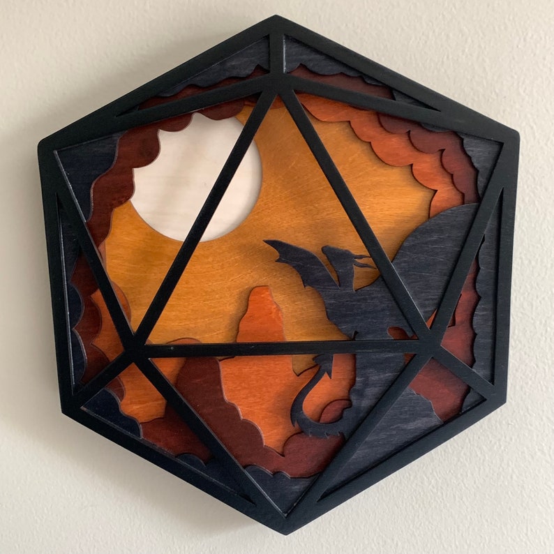 Op de afbeelding: Een zwarte houten D20 dobbelsteen met een gelaagd ontwerp met een silhouet van een draak die voor een volle maan en bergen vliegt. De dobbelsteen is gemaakt van hout en heeft een donker gebeitst oppervlak.