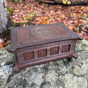 May include: A rectangular wooden box with a dark brown finish, featuring intricate engraved patterns on the lid and sides. The lid displays a central circular emblem with a triangle and smaller circles, surrounded by decorative border carvings.