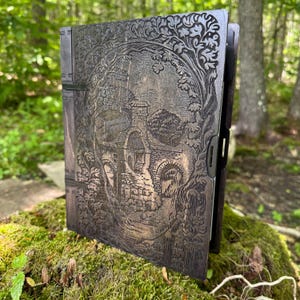 May include: A dark wooden journal with an intricate, carved design. The cover features a detailed landscape with buildings, arches, and trees, all within a skull-shaped frame. The journal is open, revealing the interior pages.