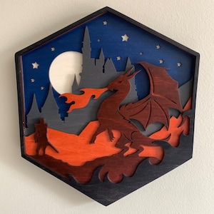 Op de afbeelding: Een houten wanddecoratie met een afbeelding van een draak die vuur spuit voor een kasteel. De draak is bruin en het kasteel is grijs. De achtergrond is donkerblauw met witte sterren. Het stuk is zeshoekig.