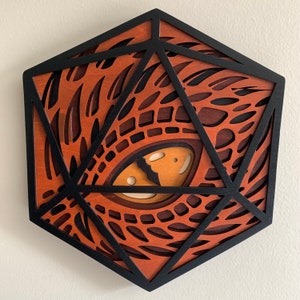 Op de afbeelding: Een geometrisch wandkunstwerk van hout in de vorm van een 20-zijdige dobbelsteen met een gedetailleerd, ingewikkeld ontwerp van een drakenoog. Het oog is gemaakt van hout en geschilderd in tinten bruin, oranje en geel.