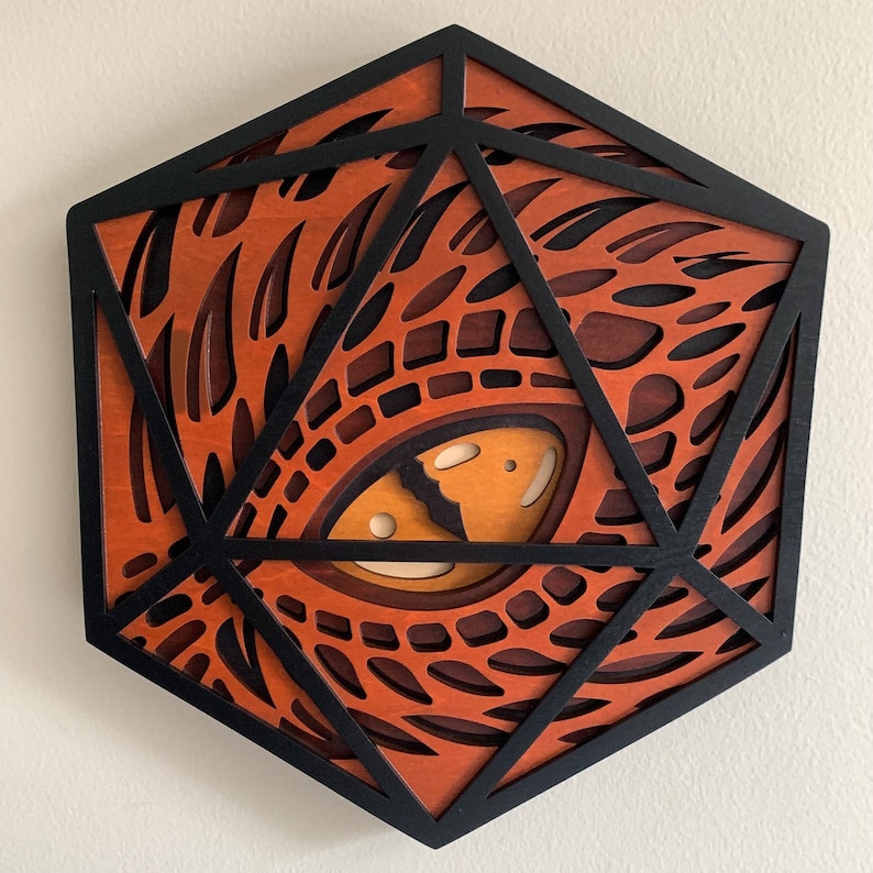 Op de afbeelding: Een geometrisch wandkunstwerk van hout in de vorm van een 20-zijdige dobbelsteen met een gedetailleerd, ingewikkeld ontwerp van een drakenoog. Het oog is gemaakt van hout en geschilderd in tinten bruin, oranje en geel.