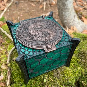 May include: A decorative wooden box with a detailed dragon design on the lid. The box is painted in shades of green and black, with a bronze-colored dragon emblem. The box has a unique scale-like pattern and is set on a bed of moss.