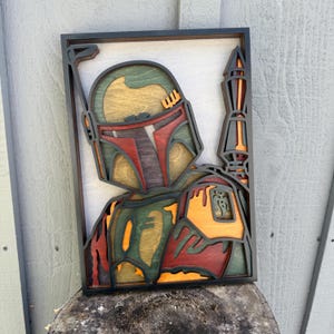 May include: A multi-layered wooden wall art piece featuring a stylized helmeted character in shades of green, red, yellow, and black. The design includes a tall antenna on the right side and a rectangular black frame, creating a dimensional effect.