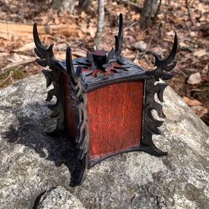 May include: A wooden box with a red stained finish and intricate black carved designs. The box has a hinged lid and a decorative metal knob.