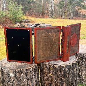 May include: A wooden, multi-panel game accessory with a red frame and intricate carvings. The panels are open, revealing a black felt interior with brass accents and a wooden interior with etched designs. The accessory is displayed on a tree stump.