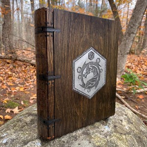 May include: A wooden book with a metal dragon design on the cover. The book has a dark brown wood finish and black metal clasps. The dragon design is in a silver color and is surrounded by a hexagonal frame.