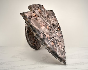 May include: A camouflage-patterned arrowhead-shaped wall mount for displaying a deer or other animal head. The mount is made of a durable material and has a realistic camouflage design.