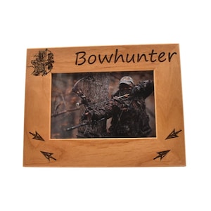 Può includere: Una cornice per foto in legno con la parola "Bowhunter" incisa nella parte superiore. La cornice contiene una foto di una persona che tiene un arco e una freccia in una zona boscosa. Ci sono frecce incise sugli angoli inferiori della cornice.