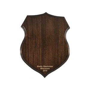 May include: A dark brown wooden shield-shaped plaque with a black border. The plaque has the text "Shelly Oberholzer Wisconsin 2019" engraved in gold lettering.