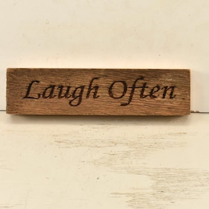 May include: A wooden sign with the text "Laugh Often" engraved in black lettering.