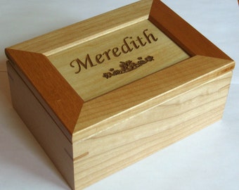 Può includere: Una scatola di legno con una finitura in legno chiaro e un bordo in legno più scuro. Il coperchio è inciso con un motivo con il nome "Meredith" e un motivo floreale.