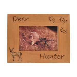Può includere: Una cornice per foto in legno con le parole "Deer Hunter" incise su di essa. La cornice ha una silhouette di cervo e corna intagliate nel legno.