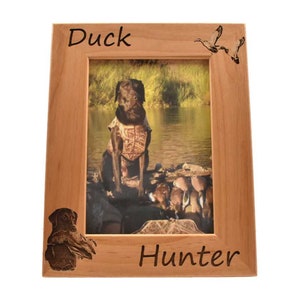 Puede incluir: Un marco de fotos de madera con una foto de un perro marr&oacute;n con un chaleco de camuflaje sentado en un bote con patos. El marco est&aacute; grabado con las palabras "Duck Hunter".