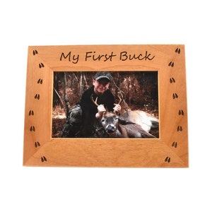 Può includere: Una cornice rettangolare in legno con la scritta "My First Buck" in nero. La cornice contiene una foto di una persona con un cervo. La cornice è decorata con impronte di cervo nere.