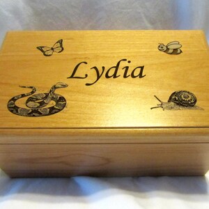 Peut inclure: Une boîte en bois avec une finition en bois clair. Le dessus de la boîte est gravé d'un papillon, d'un serpent, d'un escargot, d'une abeille et du nom "Lydia".