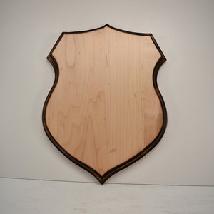 May include: A wooden shield-shaped plaque with a dark brown border. The plaque is made of light wood and has a smooth surface.