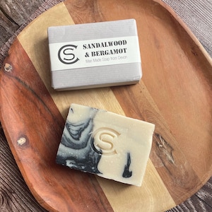 May include: Two bars of soap on a wooden tray. One is in a grey box with a label that reads "Sandalwood & Bergamot, Man Made Soap from Devon." The other bar is cream-coloured with black swirls and an embossed "S".