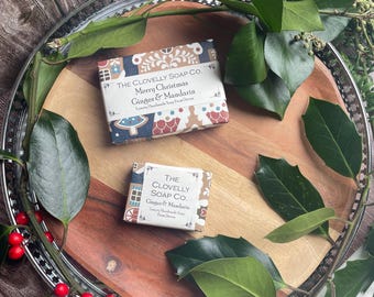 Puede incluir: Dos barras de jabón "The Clovelly Soap Co.", "Merry Christmas Ginger & Mandarin" y "Ginger & Mandarin", descansan sobre una bandeja de madera. La bandeja está rodeada de vegetación y bayas rojas, creando una exhibición festiva y de temporada. El empaque del jabón presenta un diseño navideño.