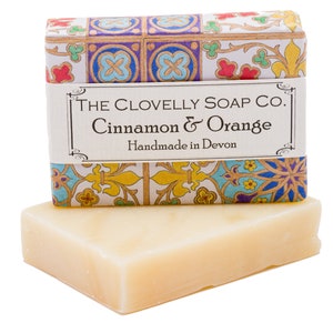 Puede incluir: Una pastilla de jabón en una caja blanca con un diseño estampado de colores. La caja tiene una etiqueta que dice "The Clovelly Soap Co. Cinnamon & Orange Handmade in Devon."