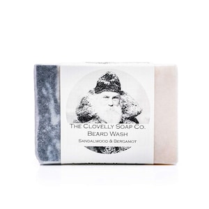 May include: A bar of soap with a black and white illustration of a man with a beard and a hat. The soap is labeled "The Clovelly Soap Co. Beard Wash Sandalwood & Bergamot"