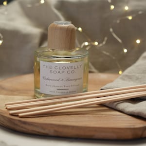 May include: A clear glass bottle with a wooden lid and label that reads "THE CLOVELLY SOAP CO. Cedarwood & Lemongrass Aromatherapy Reed Diffuser HAND POURED IN DEVON". The bottle is sitting on a wooden surface with wooden sticks.