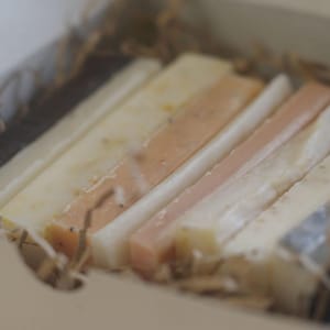 Puede incluir: Un juego de cinco barras rectangulares de jabón en varios tonos de blanco, beige y rosa. Las barras de jabón están dispuestas en una caja con papel triturado.