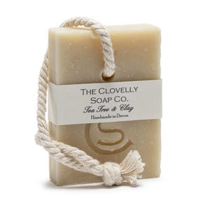 Op de afbeelding: Een beige zeep met een touw eraan bevestigd. De zeep heeft de tekst "THE CLOVELLY SOAP CO. Tea Tree & Clay Handmade in Devon" op een etiket.