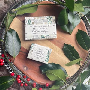 Puede incluir: Dos jabones navideños con la inscripción "Merry Christmas Oh Christmas Tree!" y un diseño de pueblo. Se presentan en una bandeja de madera, rodeados de vegetación y bayas rojas, creando un ambiente festivo. Jabones artesanales.
