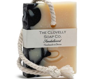 May include: A bar of handmade sandalwood soap with a black and white marbled design. The soap is attached to a white rope with a knot at the end. The soap has a label that reads "The Clovelly Soap Co. Sandalwood Handmade in Devon."