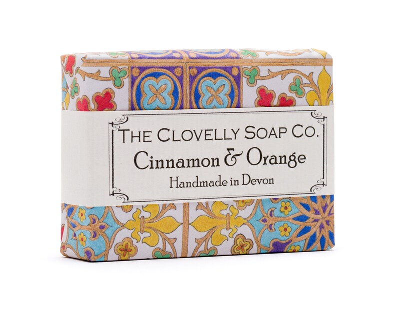 May include: A bar of soap with a colourful, patterned wrapper. The wrapper features a label that reads "THE CLOVELLY SOAP CO. Cinnamon & Orange Handmade in Devon."