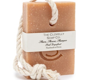 Puede incluir: Una pastilla de jabón de pomelo rosa con una cuerda para colgar. El jabón está etiquetado con "The Clovelly Soap Co. Shave, Shower, Shampoo Pink Grapefruit Handmade in Devon."