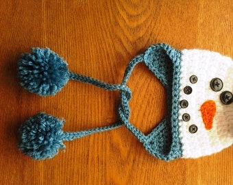 Puede incluir: Sombrero de punto blanco de muñeco de nieve con sombrero de copa negro, nariz naranja y ojos de botón negro. El sombrero tiene orejeras azules con pompones azules en los extremos.