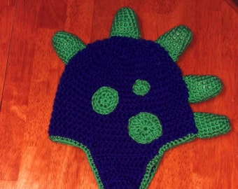 May include: A blue and green crocheted hat with a dinosaur design. The hat has a long, braided cord.
