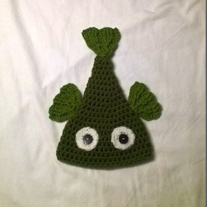 May include: A green crocheted fish hat with white button eyes. The hat has a triangular shape with a pointed top and a tail fin.