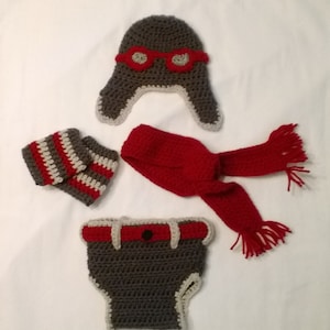 May include: A crocheted baby outfit set in gray and red. The set includes a hat with red goggles, a scarf, booties, and a diaper cover.