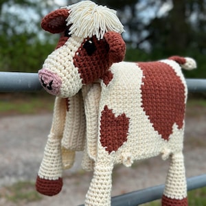 May include: A crocheted cow-shaped baby blanket with brown and white spots. The blanket is hanging on a metal fence.
