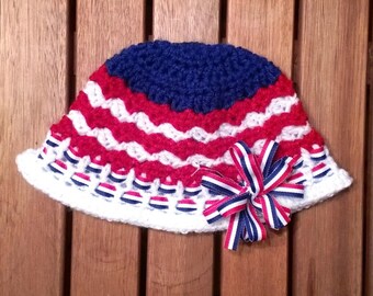 May include: A white crocheted hat with red, white, and blue stripes and a blue crocheted top. A red, white, and blue ribbon bow is attached to the hat.
