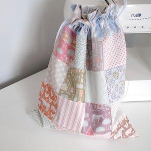 Peut inclure: Un sac à cordon patchwork aux couleurs pastel, notamment rose, bleu et beige. Le sac est composé de divers carrés de tissu avec des motifs tels que des rayures, des pois et de petites illustrations. Le sac est posé sur une surface blanche.
