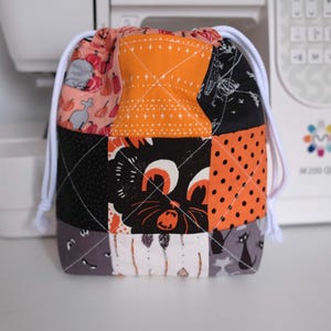 May include: A patchwork drawstring bag with a Halloween theme. The bag features squares of orange, black, and grey fabric with Halloween-themed patterns, including a black cat, pumpkins, and polka dots. White drawstring closure.