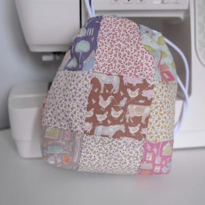 Op de afbeelding: Een patchwork-trekkoordtas met diverse stofpatronen, waaronder bloemen, dieren en geometrische ontwerpen. De tas is gemaakt van aan elkaar genaaide stofvierkanten. Een wit trekkoord is zichtbaar aan de bovenkant.