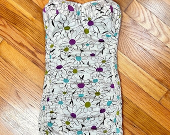 Può includere: Un costume da bagno bianco con scollo a halter e stampa floreale con contorni neri, dettagli viola, verdi e blu. Il costume da bagno ha un corpetto arricciato e una silhouette aderente.